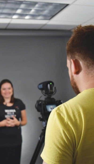 A person in a yellow shirt films another person in a grey room. The person is using a tripod-mounted camera.