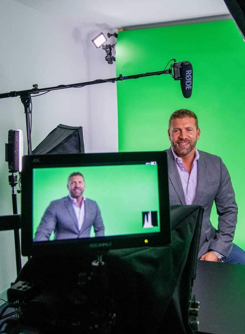A person in a gray suit is recorded in a studio with a green screen and professional lighting setup visible in the background.