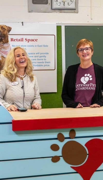 Two people stand smiling behind a reception counter in a pet retail space, featuring colorful decor and promotional signage.