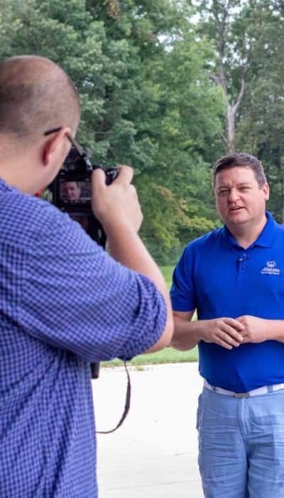 Two people outdoors, one holding a camera capturing the other who is speaking. Green foliage and trees are in the background.