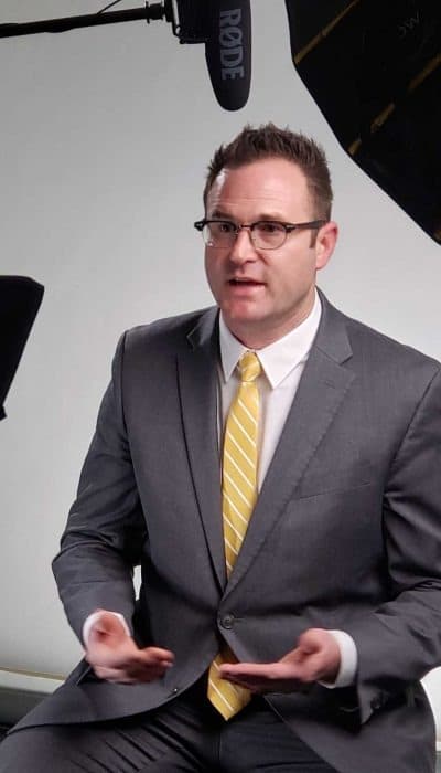 A person in a suit and yellow tie sits in a studio with professional lighting and microphone equipment, appearing engaged in conversation.
