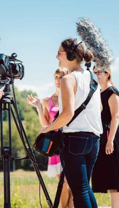 Three people are outdoors, filming with professional equipment. Microphone operator holds boom mic. Sunny day with trees in the background.